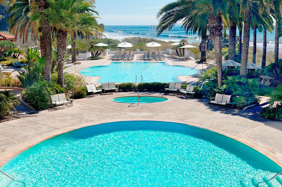 Resort pool complex with multiple turquoise pools, white loungers, palm trees, and ocean view beyond
