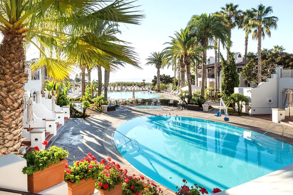 Resort pool area with palm trees, white architecture, and mountain views in background