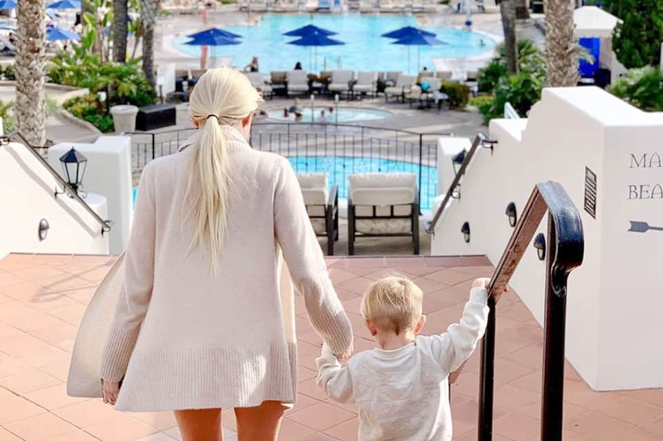 Woman and child walking hand-in-hand down terracotta stairs toward resort pools and palm trees