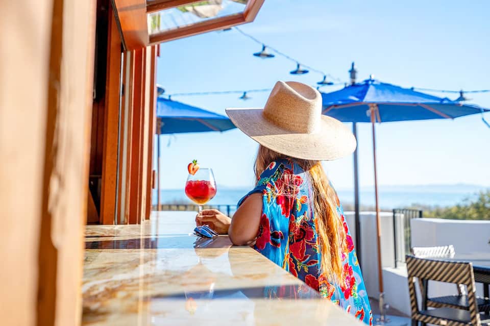 Woman in colorful dress and straw hat enjoying red cocktail at oceanview bar with blue umbrellas visible in background