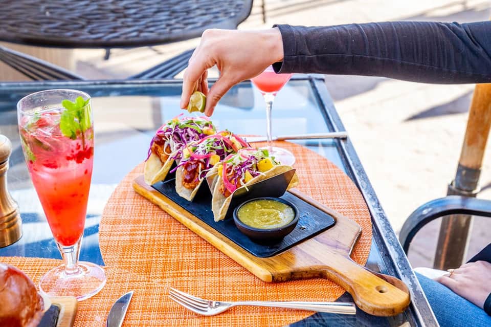 Beachside dining table with tacos, cocktails, and person reaching for lime garnish