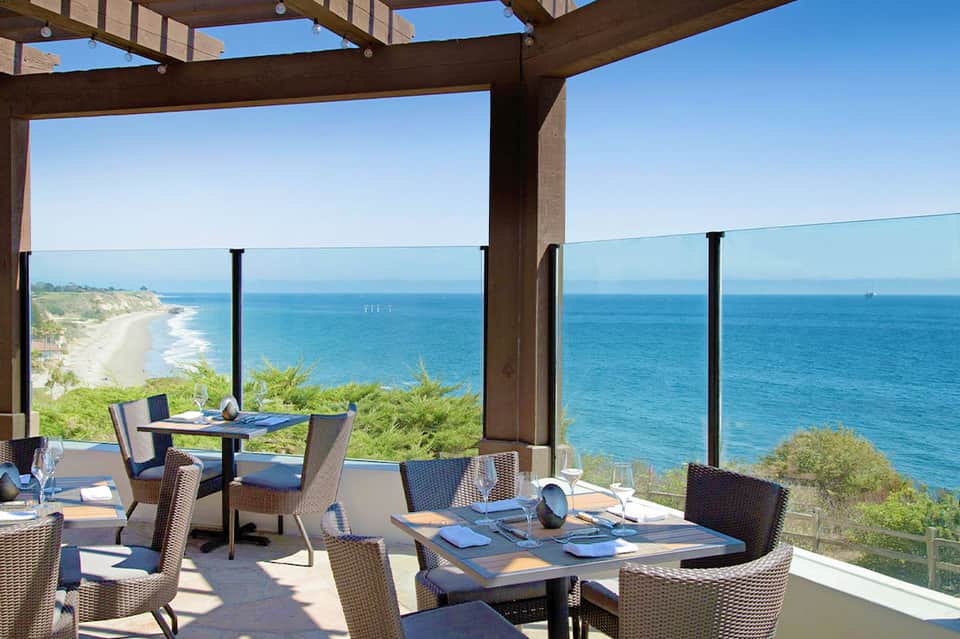Coastal restaurant terrace with ocean and beach views, wooden pergola, modern furniture overlooking cliffs