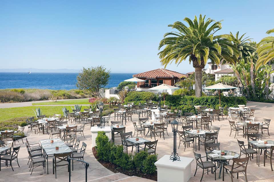 Oceanfront patio dining area with tables, chairs, palms, and Mediterranean-style villa at sunset