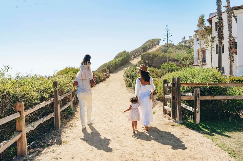 Family walking on sandy beach path with wooden railings toward ocean, white coastal buildings