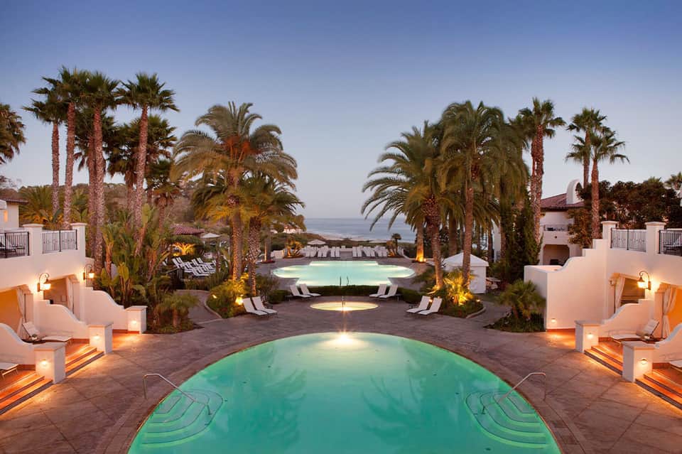 Illuminated resort pools at dusk surrounded by palm trees, loungers, and white buildings