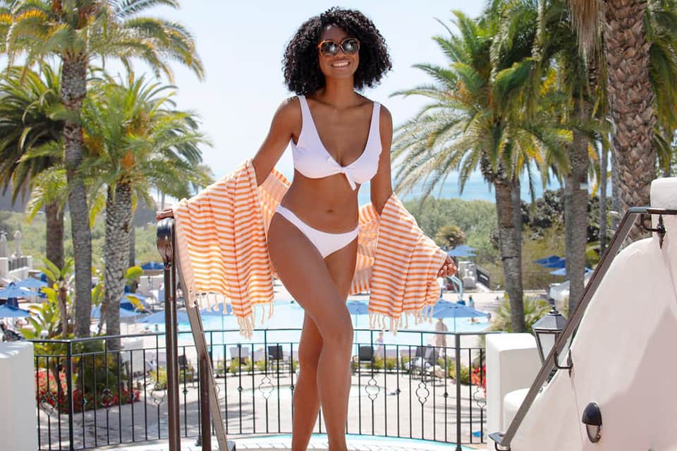 Woman in white bikini and orange striped cover-up on resort balcony overlooking pool and palm trees