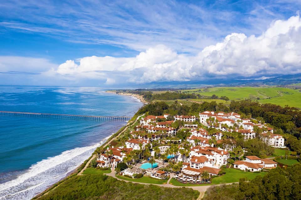 Aerial view of beachfront residential community with white buildings, pools, and green coastal cliffs