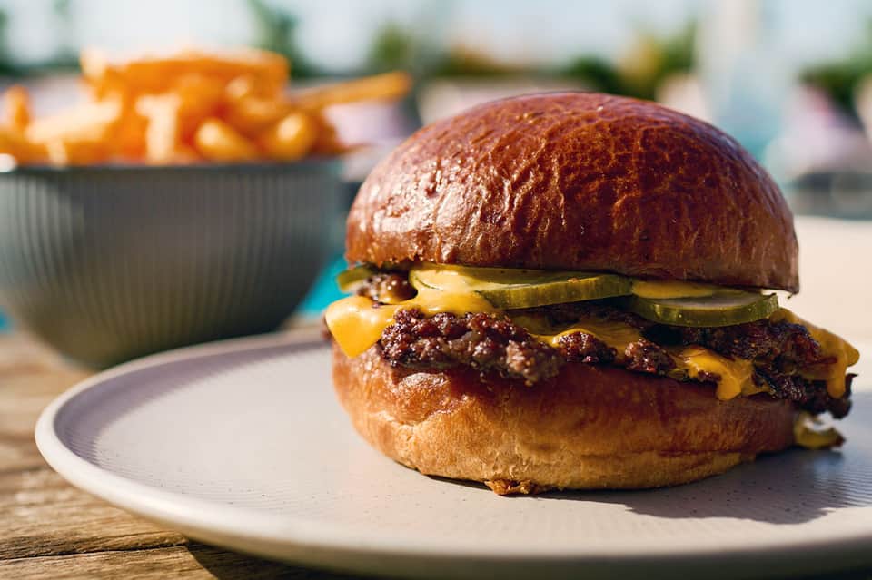 Gourmet burger with cheese and pickles served on white plate with side dish outdoors