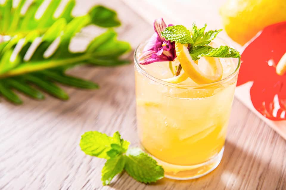 Yellow cocktail with lemon twist, mint leaves, and purple flower garnish on wooden surface