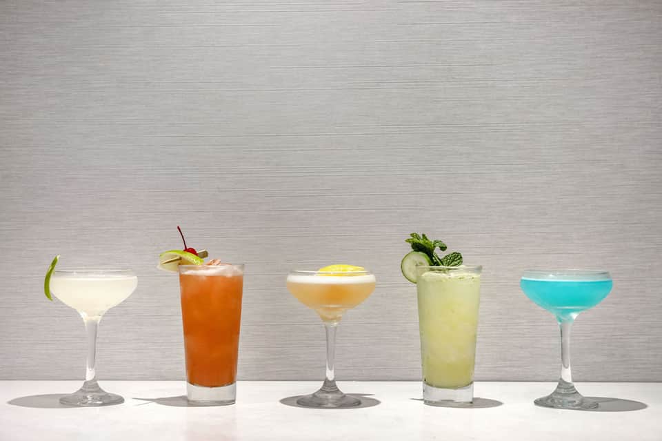 Five tropical cocktails in various colors displayed on white surface with garnishes