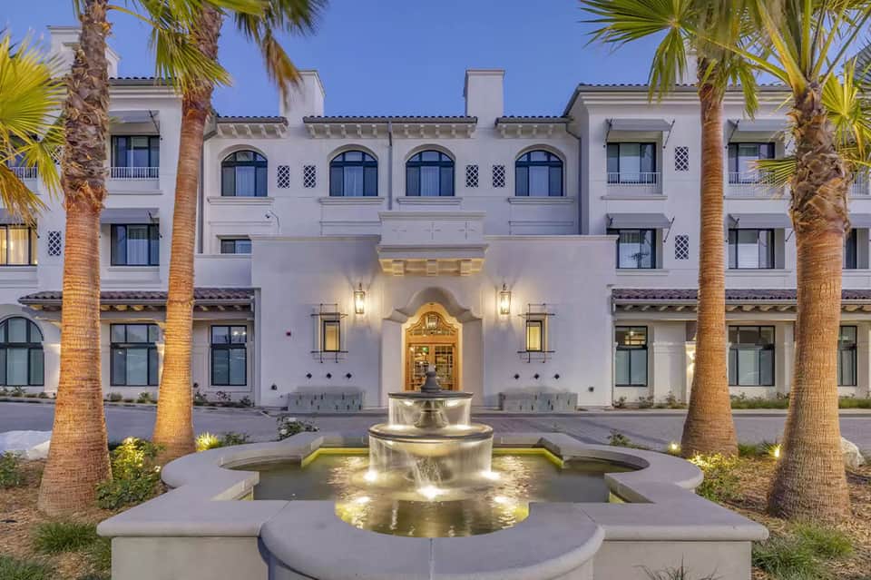 Illuminated Mediterranean-style white resort building courtyard with fountain at dusk surrounded by palms
