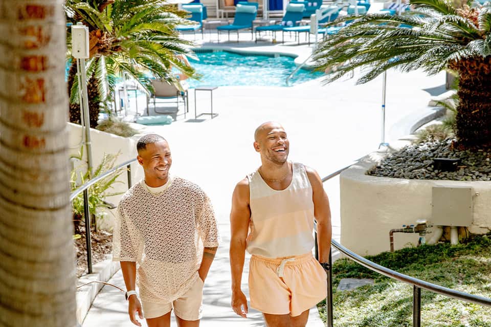 Two men walking down a sunny pathway toward a pool area with palm trees and resort buildings