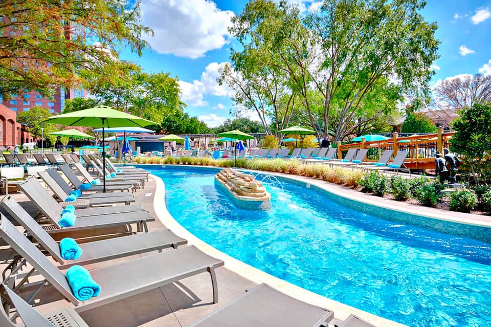Bright outdoor pool with lounge chairs, green umbrellas, and lush landscaping under blue sky