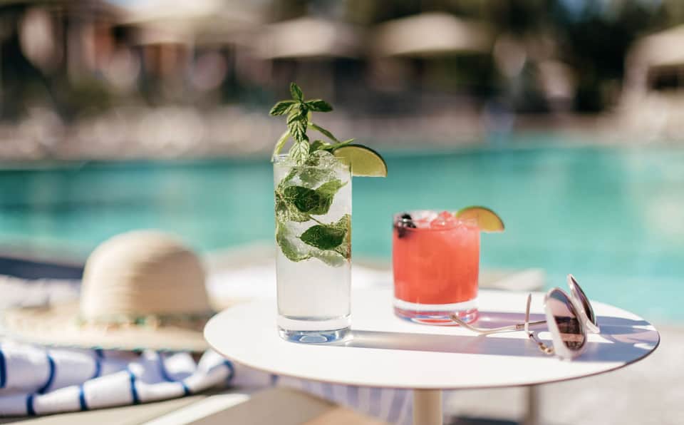 Two refreshing cocktails on poolside table with hat, sunglasses, and blurred resort background