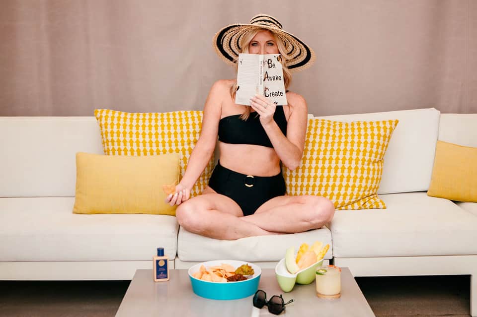Woman in bikini on couch with yellow pillows holding notebook near snacks