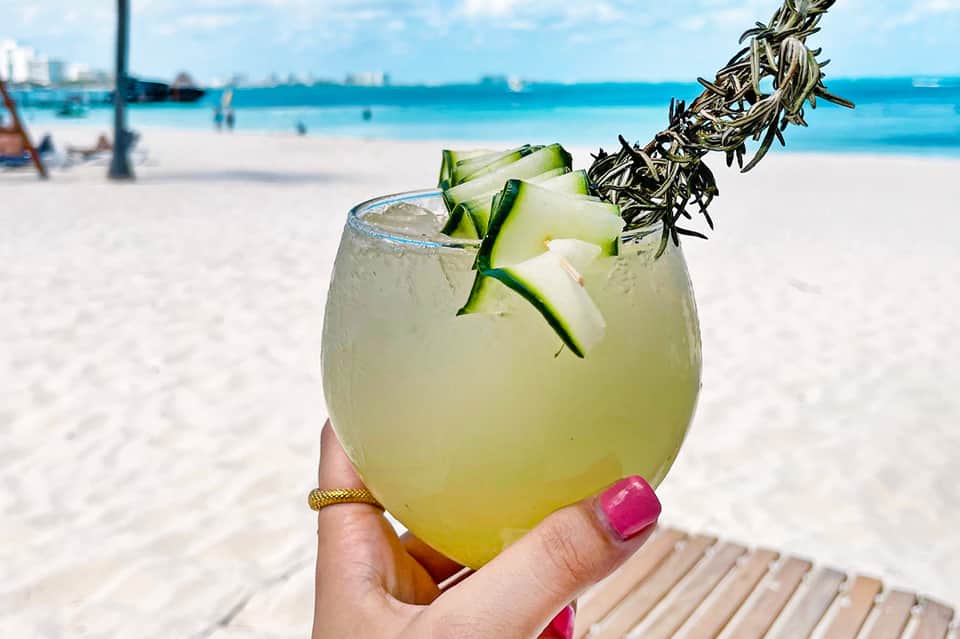 Tropical pineapple cocktail with cucumber garnish held at beach with ocean in background