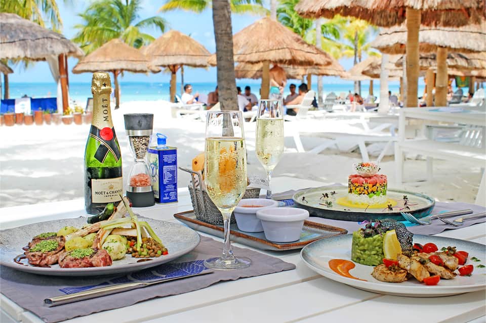 Beachfront dining table with champagne, gourmet plates, and thatched umbrellas