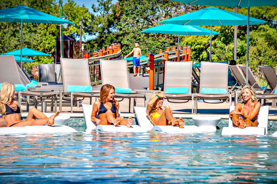 Adults relaxing in pool loungers under turquoise umbrellas at resort pool with playground visible