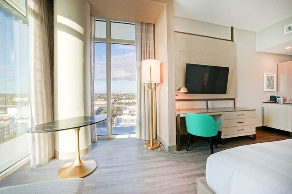 Luxury hotel room with floor-to-ceiling window view, turquoise chair at desk, modern furnishings and natural light.