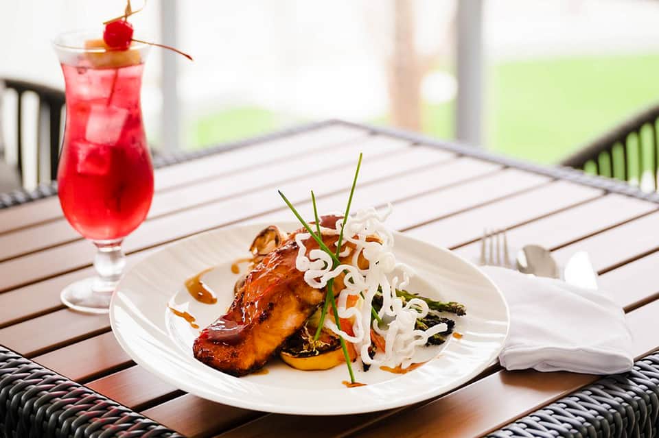 Plated grilled fish dish with red cocktail on outdoor dining deck with green lawn view