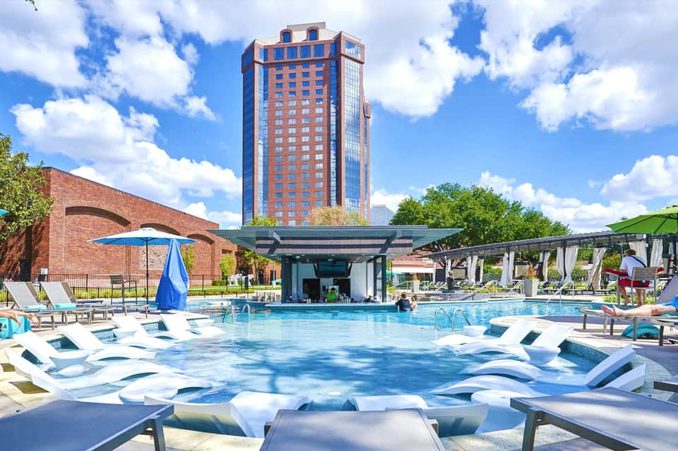 Outdoor resort pool with lounge chairs, red brick building, pergola, and blue umbrellas under clear sky