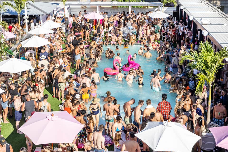 Crowded pool party with hundreds of guests, pink floats, white umbrellas, and tropical setting