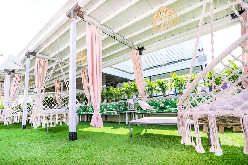 Outdoor event space with white pergola, pink draped fabric, artificial turf, and tropical plants