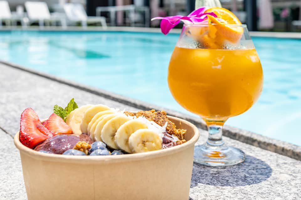Tropical smoothie bowl and orange cocktail poolside with ocean view in background