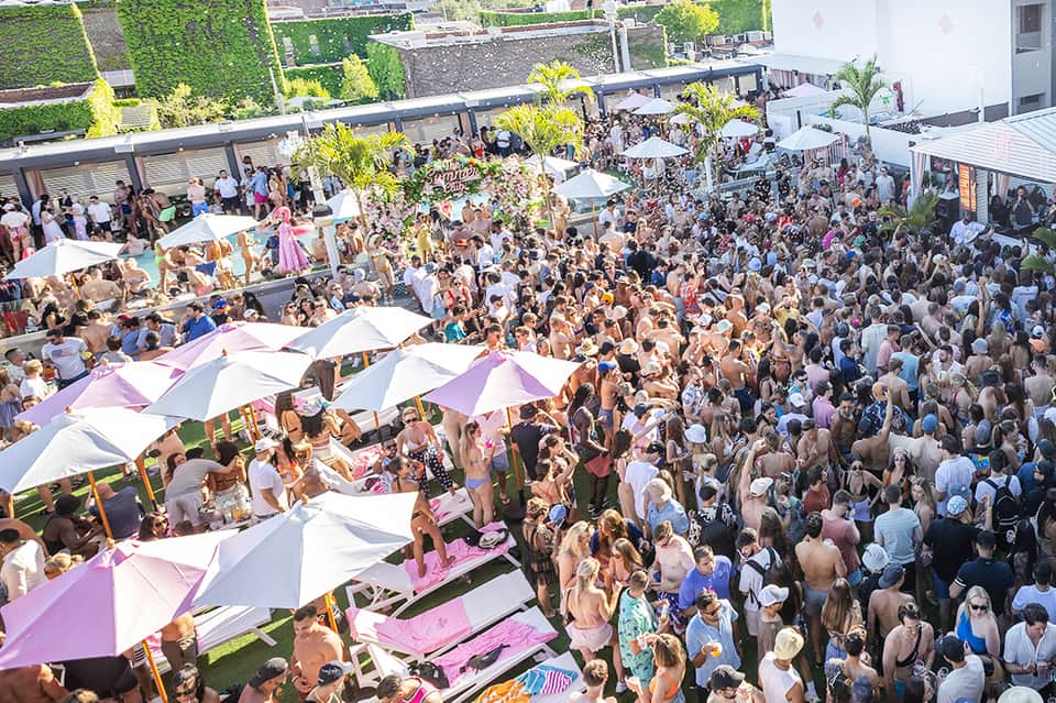 Aerial view of crowded rooftop pool party with colorful umbrellas, loungers, and hundreds of guests
