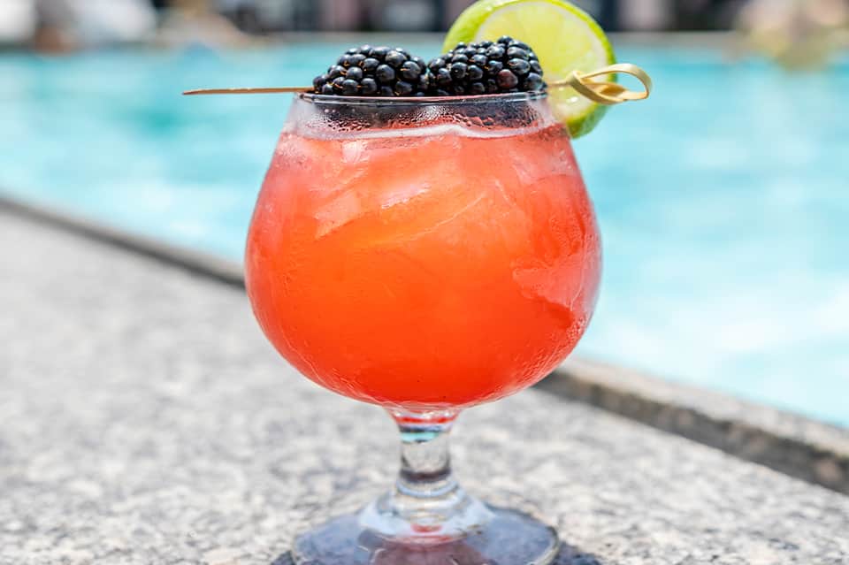 Coral-colored tropical cocktail with blackberries and lime wheel by swimming pool