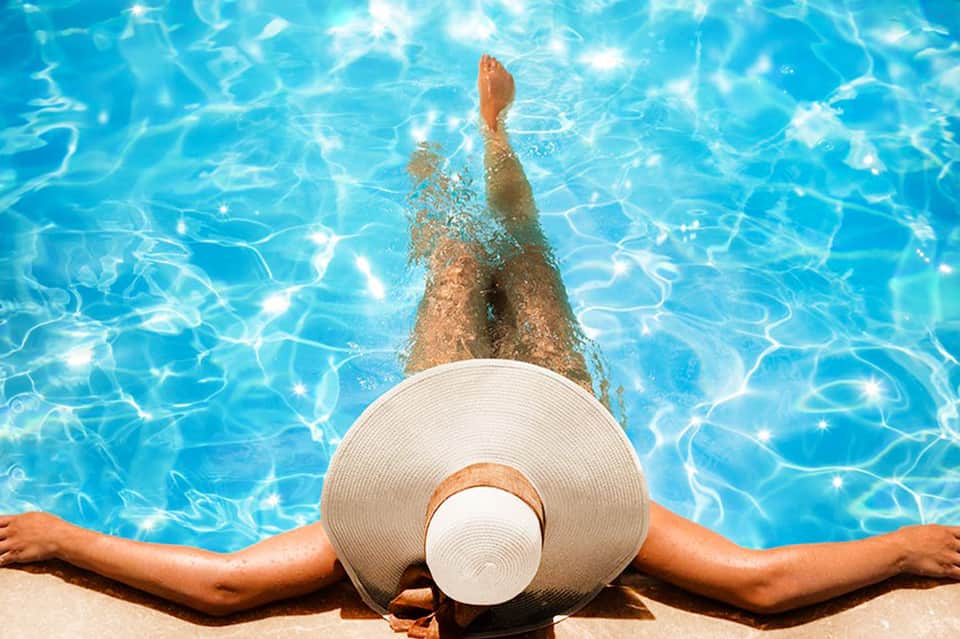Person floating in clear blue pool wearing white sun hat, viewed from above
