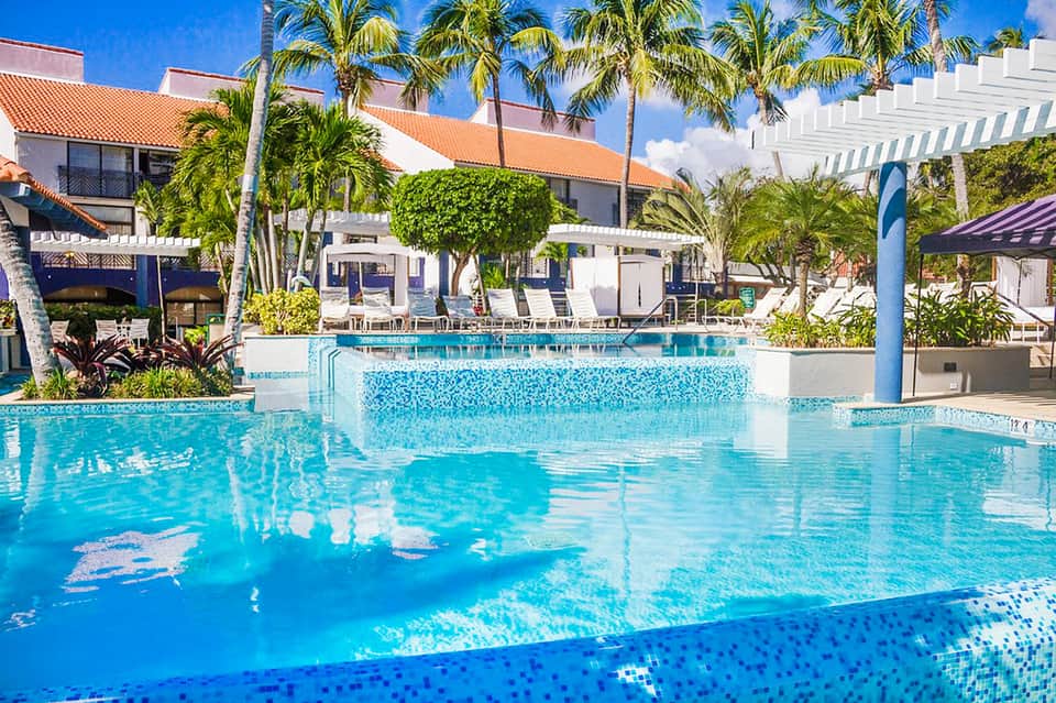 Resort swimming pool with palm trees, white buildings, and lounge chairs under blue sky