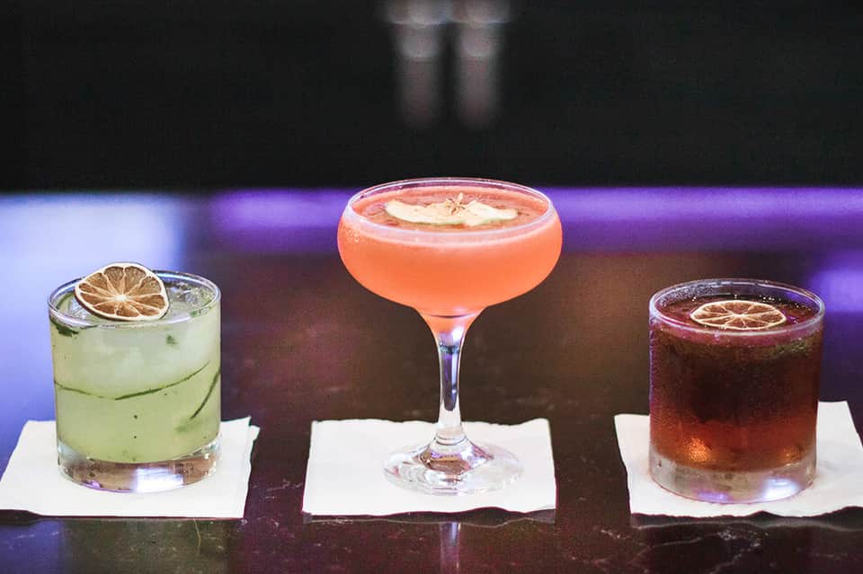 Three colorful cocktails with citrus garnishes displayed at resort bar with purple lighting