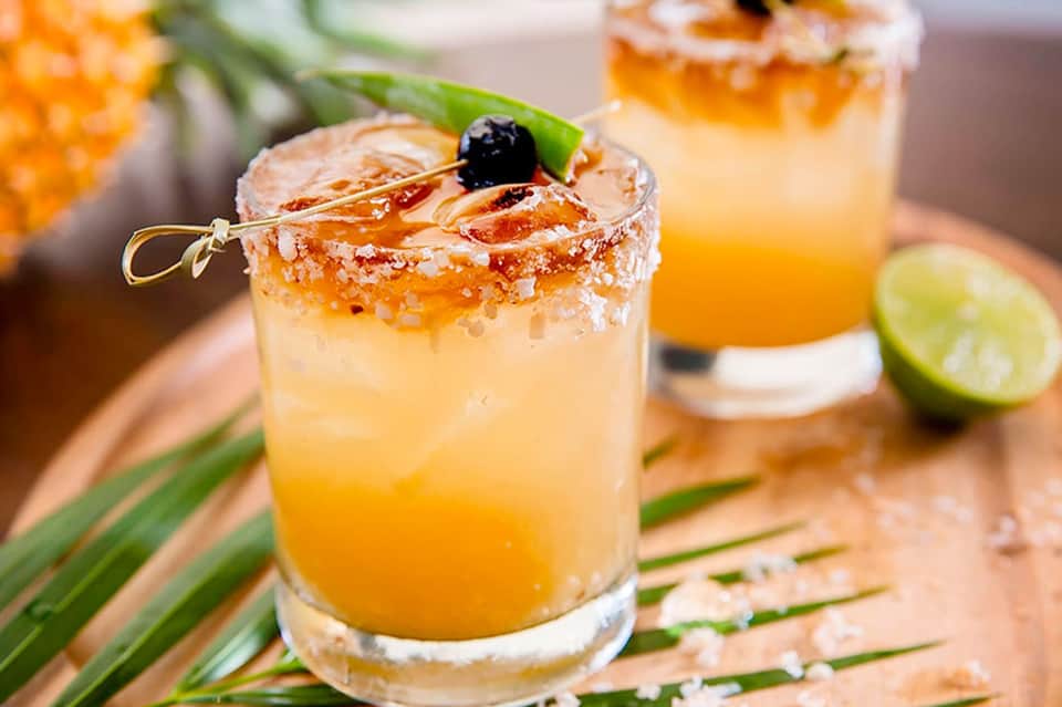 Tropical cocktail with spiced rim, black olive garnish, and lime wedge on wooden surface