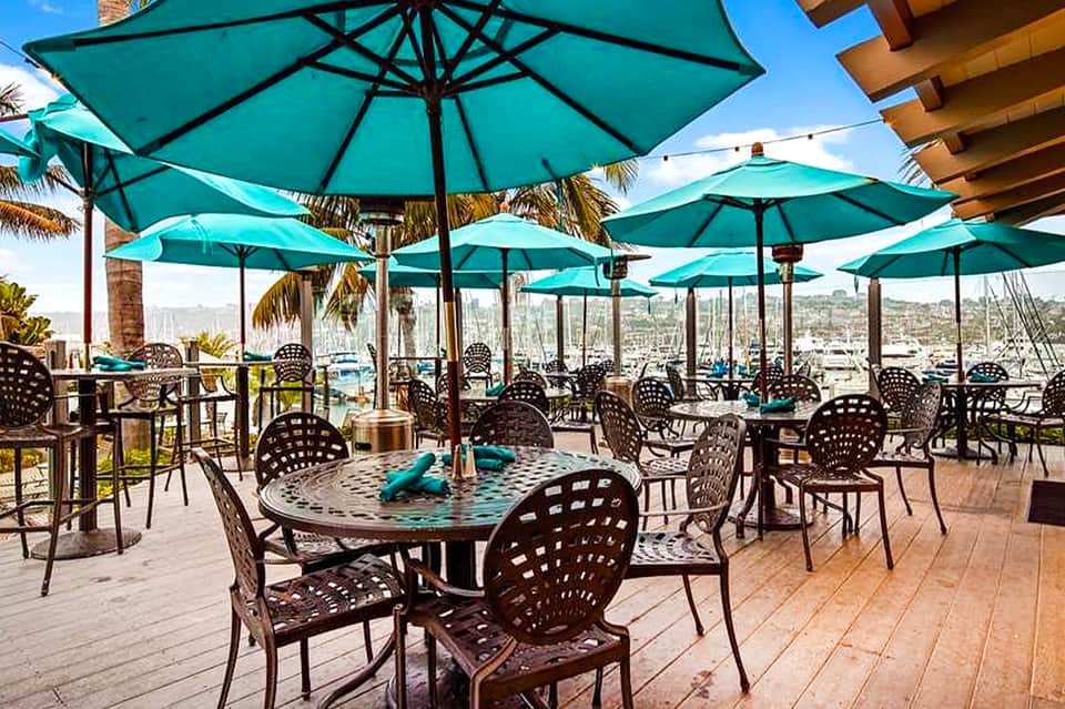 Waterfront dining area with turquoise umbrellas, cast iron furniture, wooden deck, and harbor views