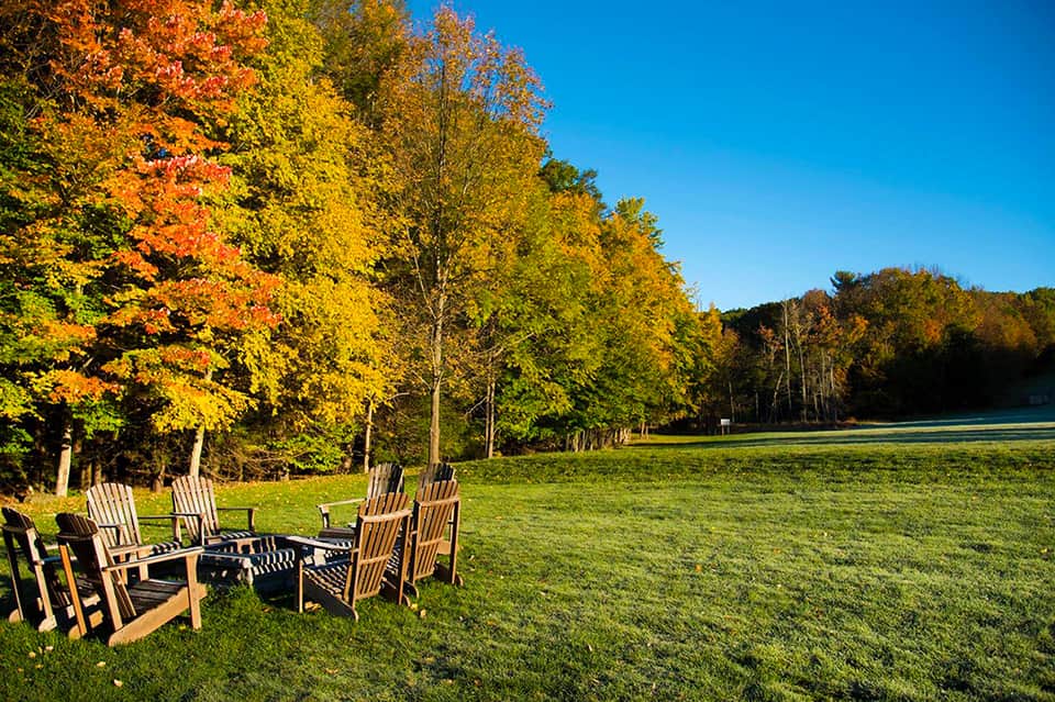 Wooden Adirondack chairs by a lakeside fire pit with autumn foliage trees