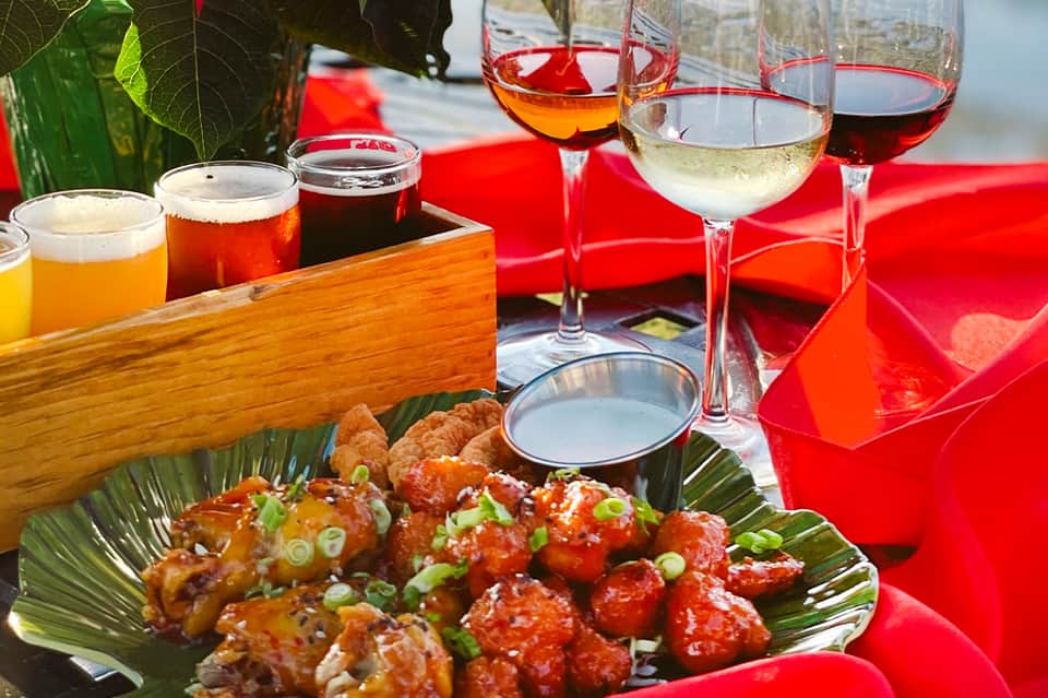 Outdoor dining setup with various cocktails, beer, wine glasses, and Asian appetizers on red tablecloth