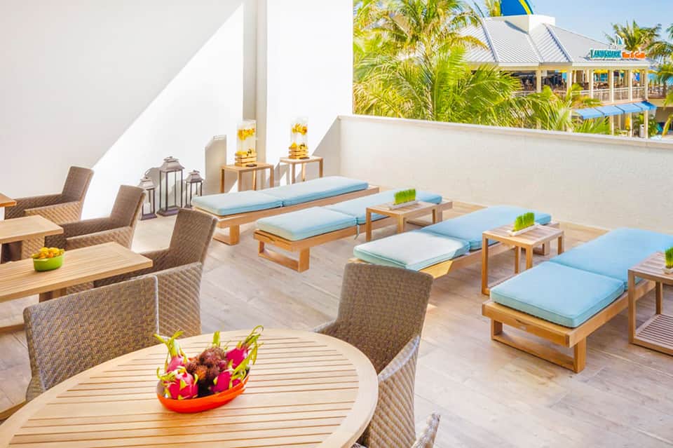 Tropical rooftop terrace with wooden lounges, blue cushions, and palm trees overlooking a beachfront resort