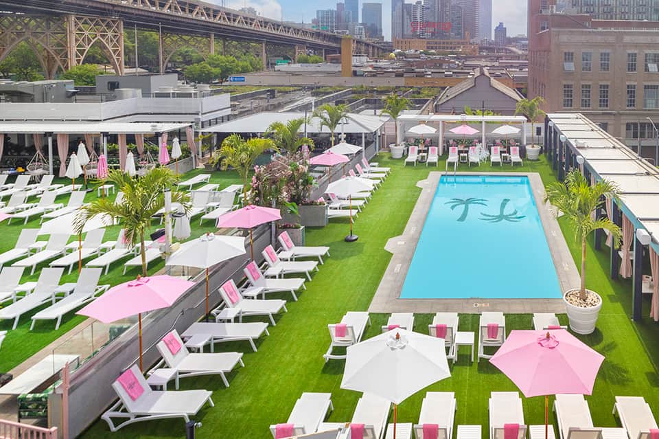 Rooftop pool deck with pink and white umbrellas, lounge chairs, and city skyline with bridge view