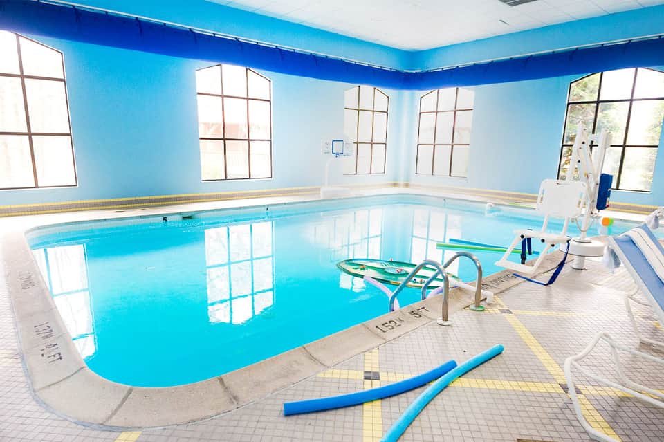 Indoor swimming pool with bright blue water, windows, and exercise equipment