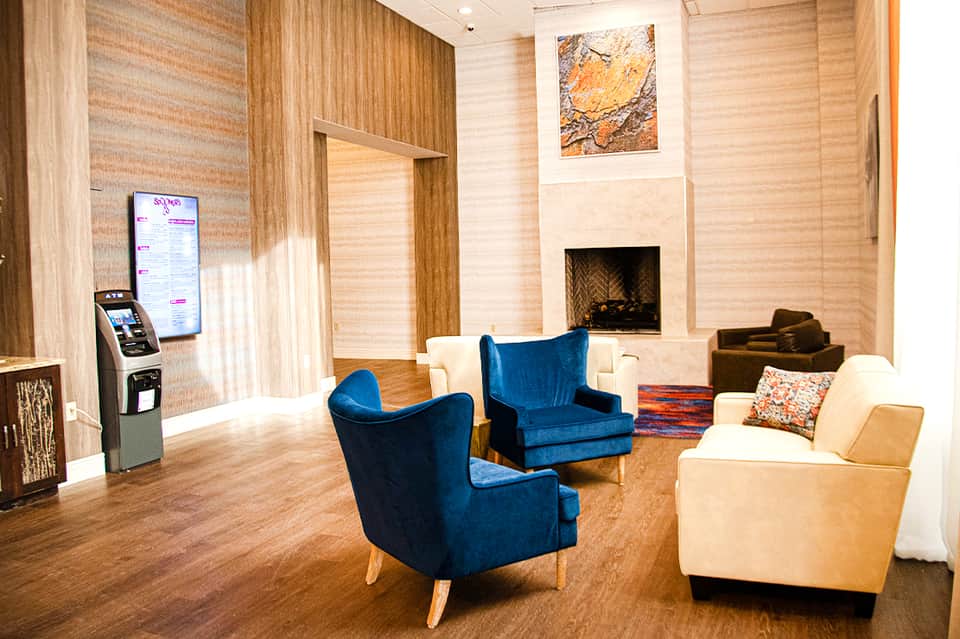 Contemporary hotel lobby with blue accent chairs, fireplace, wood paneling, and ATM kiosk