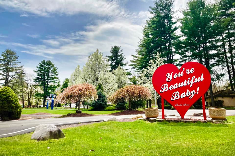 Red heart-shaped 'You're Beautiful Baby' sign at a resort entrance with trees and green lawn