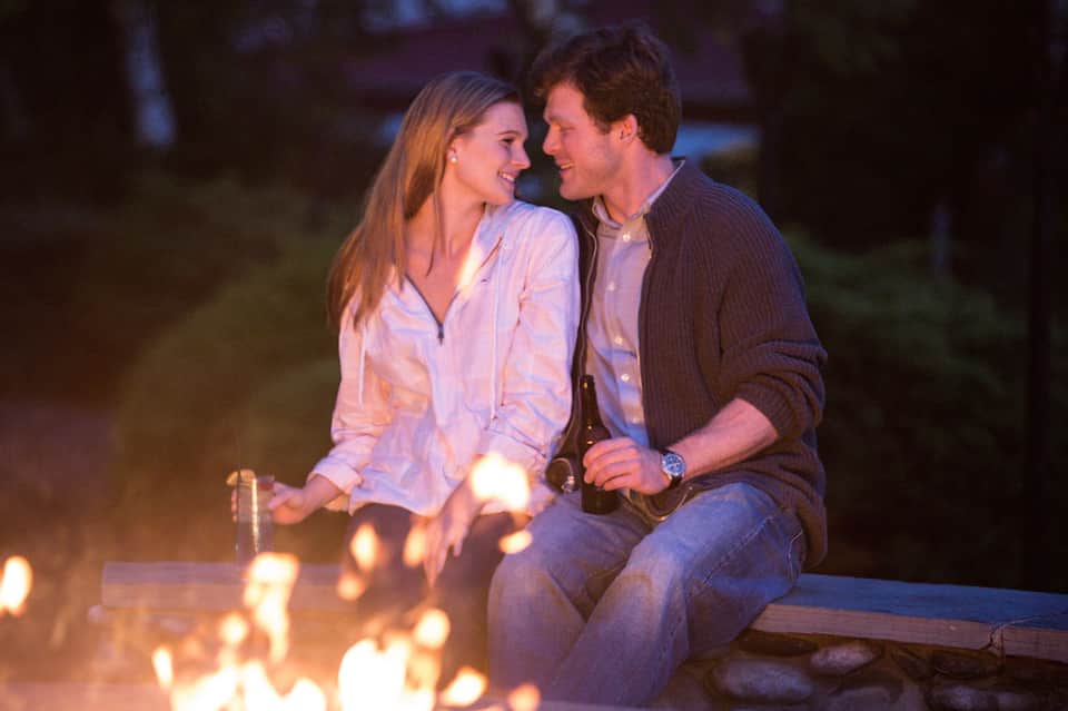 Couple sitting by campfire at dusk holding beer bottle and smiling
