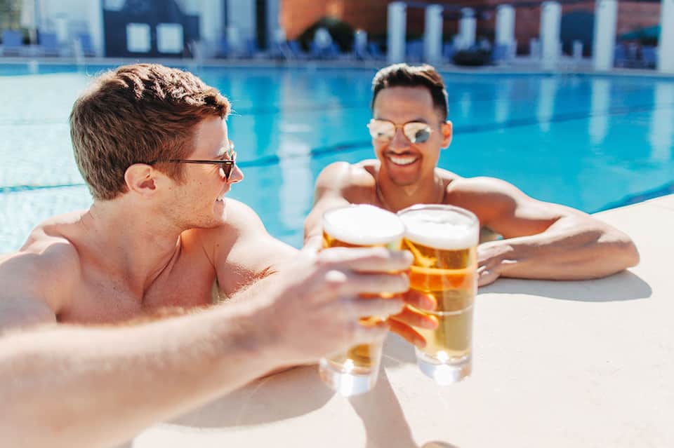 Two men relaxing in pool holding beer glasses with blue water backdrop