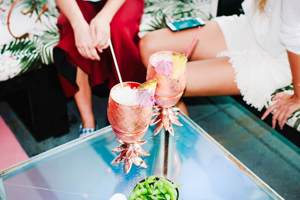 Two tropical cocktails with pink umbrellas held by people in festive attire