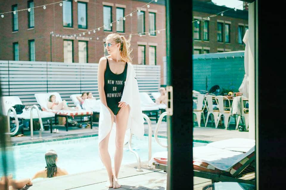 Woman in black swimsuit standing by rooftop pool with brick buildings, string lights, and other guests in background