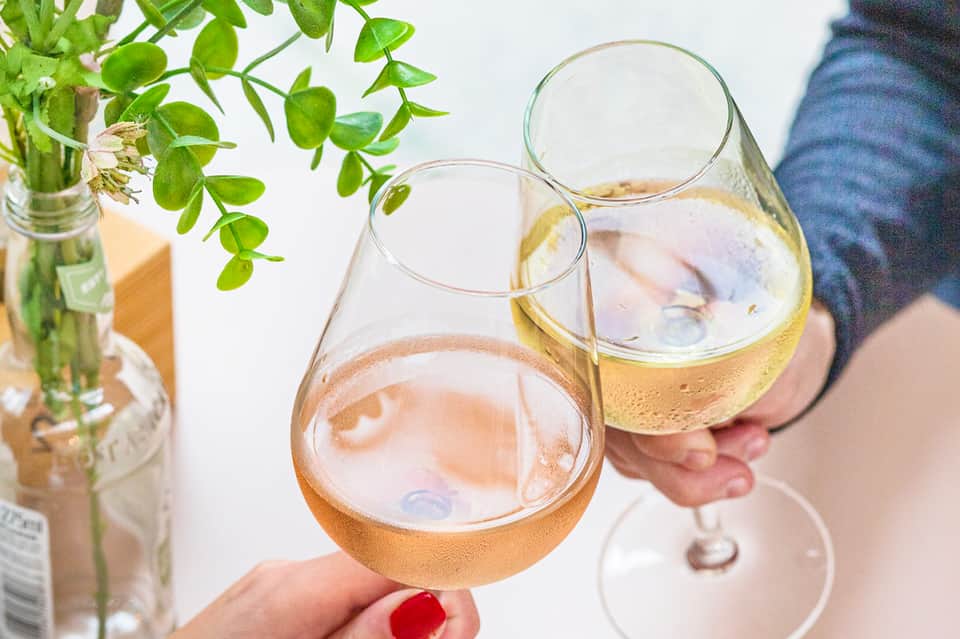 Two hands holding wine glasses with rosé and white wine outdoors near green foliage