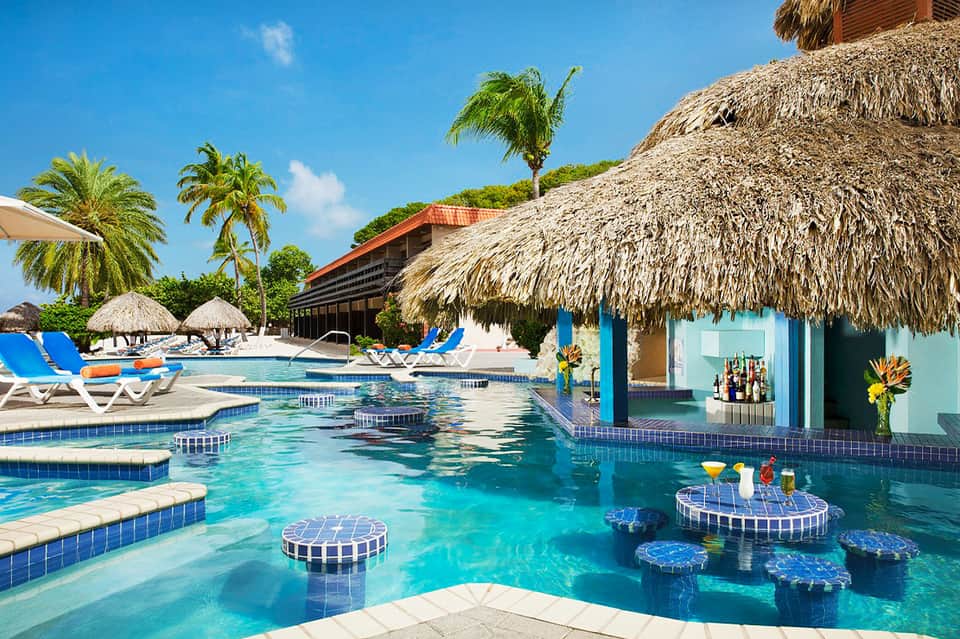 Resort pool with thatched roof cabanas, blue loungers, and tropical landscaping overlooking water