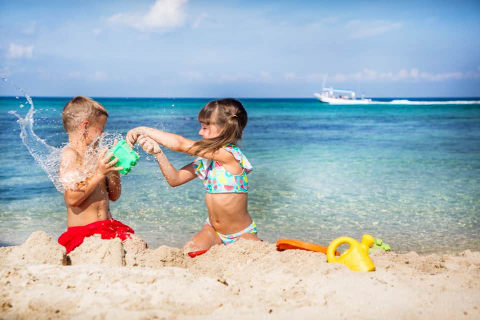 Two children playing in shallow ocean water with colorful beach toys and clear blue sea