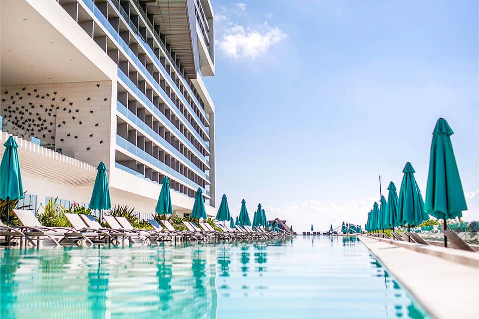 Beachfront resort pool with turquoise water, lounge chairs, green umbrellas, and modern building with balconies
